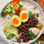 A Korean bbq rice bowl fully assembled with a gold fork, kitchen towel, and small dish of carrots next to it.