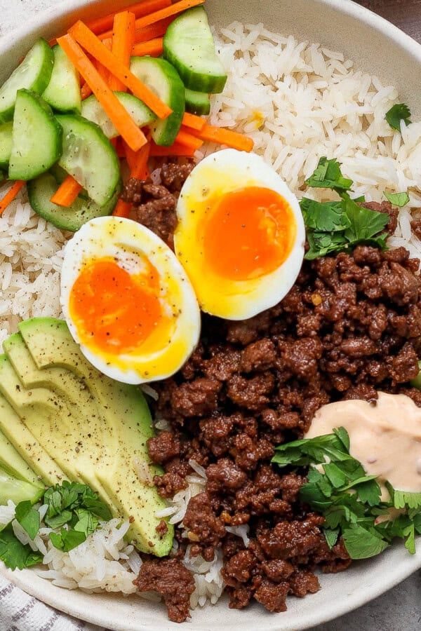 A Korean bbq rice bowl fully assembled with a gold fork, kitchen towel, and small dish of carrots next to it.