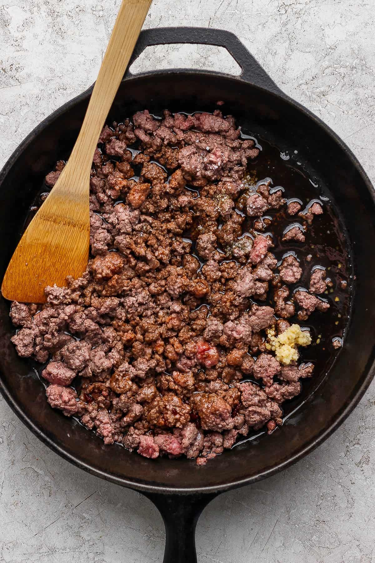 Browned ground beef in a cast iron skillet with a wooden spoon and minced garlic, salt, pepper, and Korean BBQ sauce added to it.