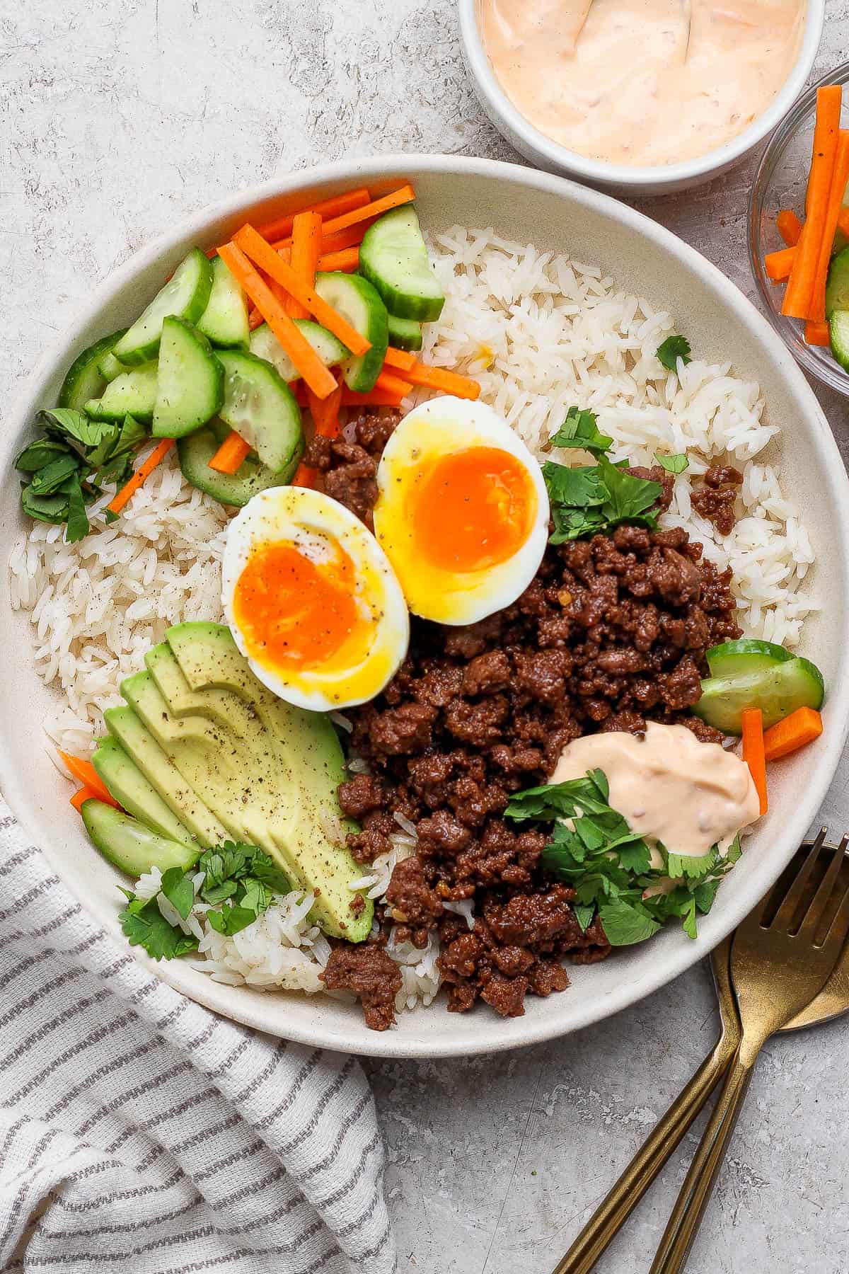 A fully assembled Korean BBQ bowl next to gold silverware, a kitchen towel, bowl of chili garlic mayo, and another bowl of the salad.