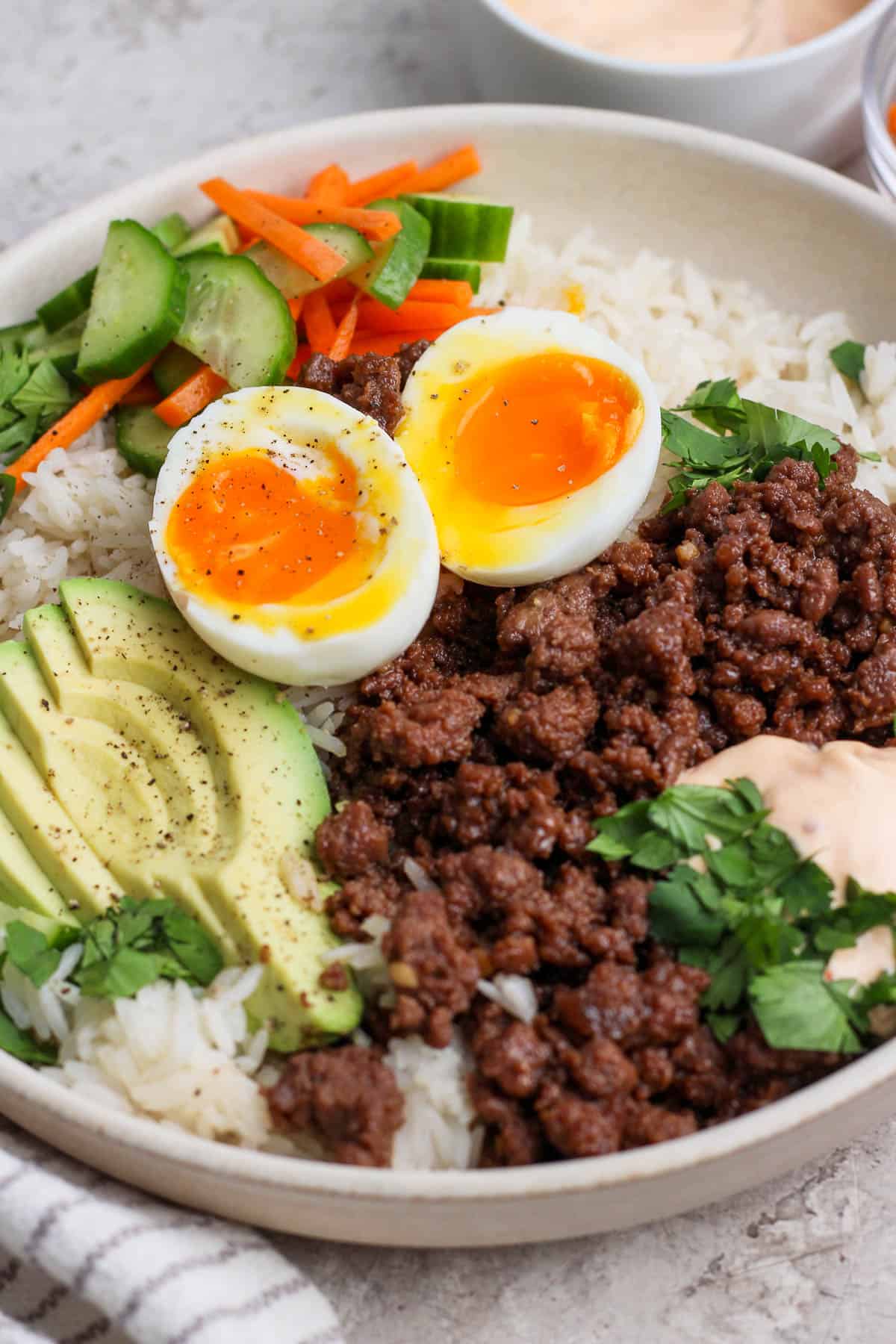 A Korean BBQ bowl with a soft boiled egg on top, fresh cilantro, avocado slices, salad, and chili garlic mayo.