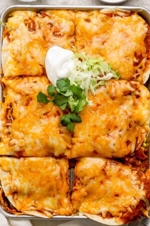 A pan of sheet pan enchiladas with shredded beef, cut into squares and topped with sour cream, shredded lettuce and chilantro.