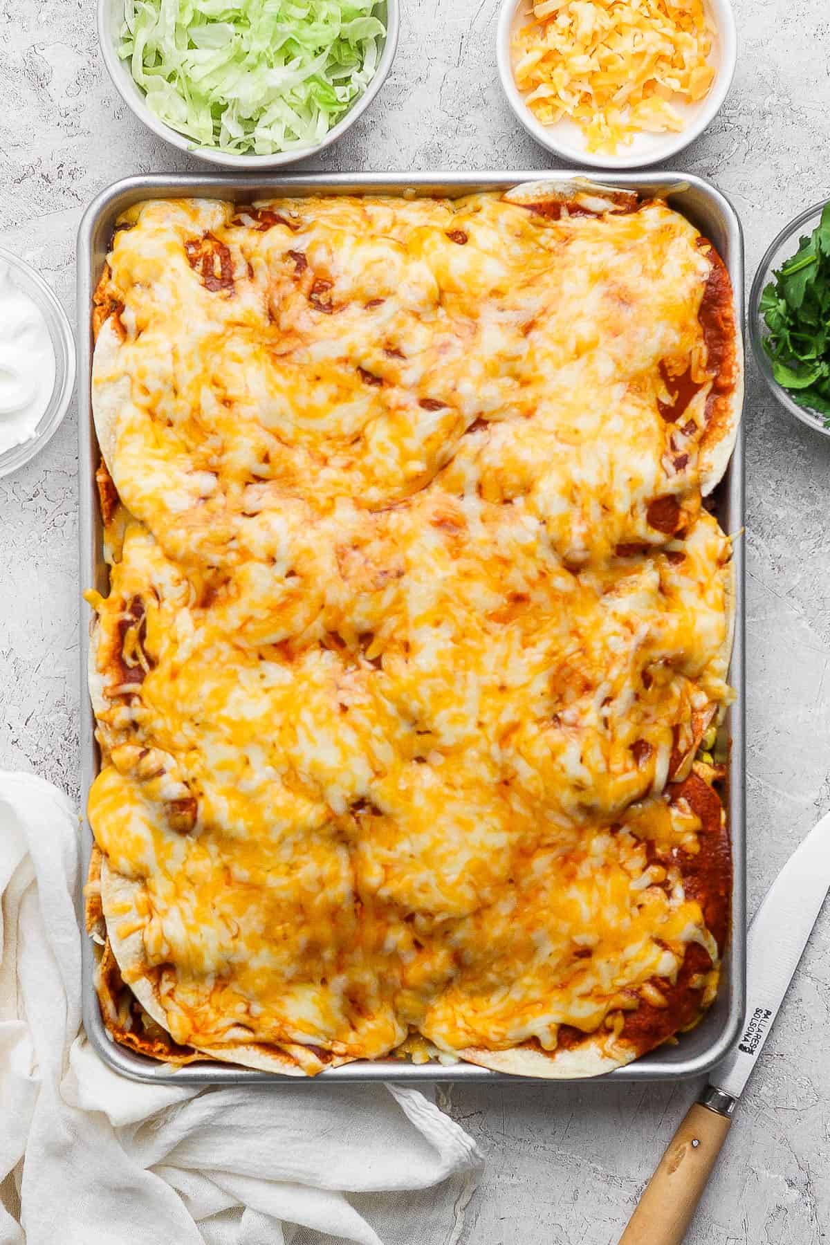 Sheet pan enchiladas after baking in the oven with small bowls of cheese, lettuce, sour cream, and cilantro around it.