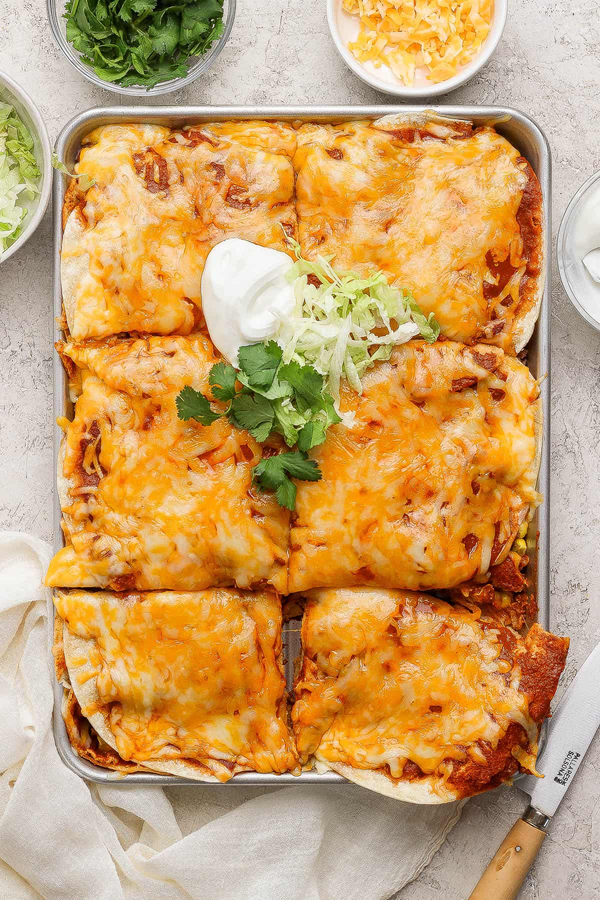 A pan of sheet pan enchiladas after baking with some lettuce and sour cream added on top next to bowls of cheese, lettuce, and cilantro.