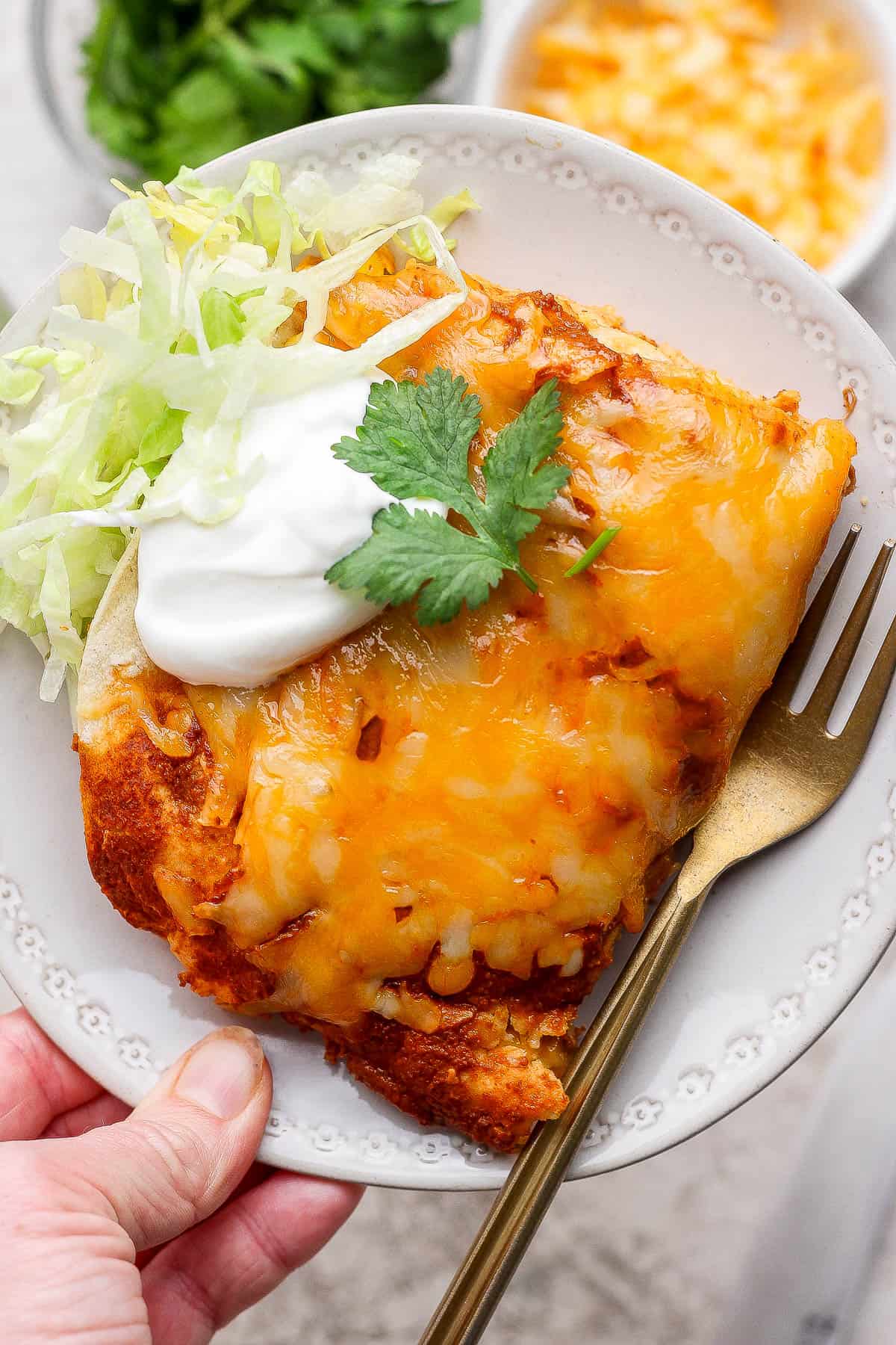 A serving of sheet pan enchiladas on a plate with some shredded lettuce, sour cream, cilantro, and a gold fork.