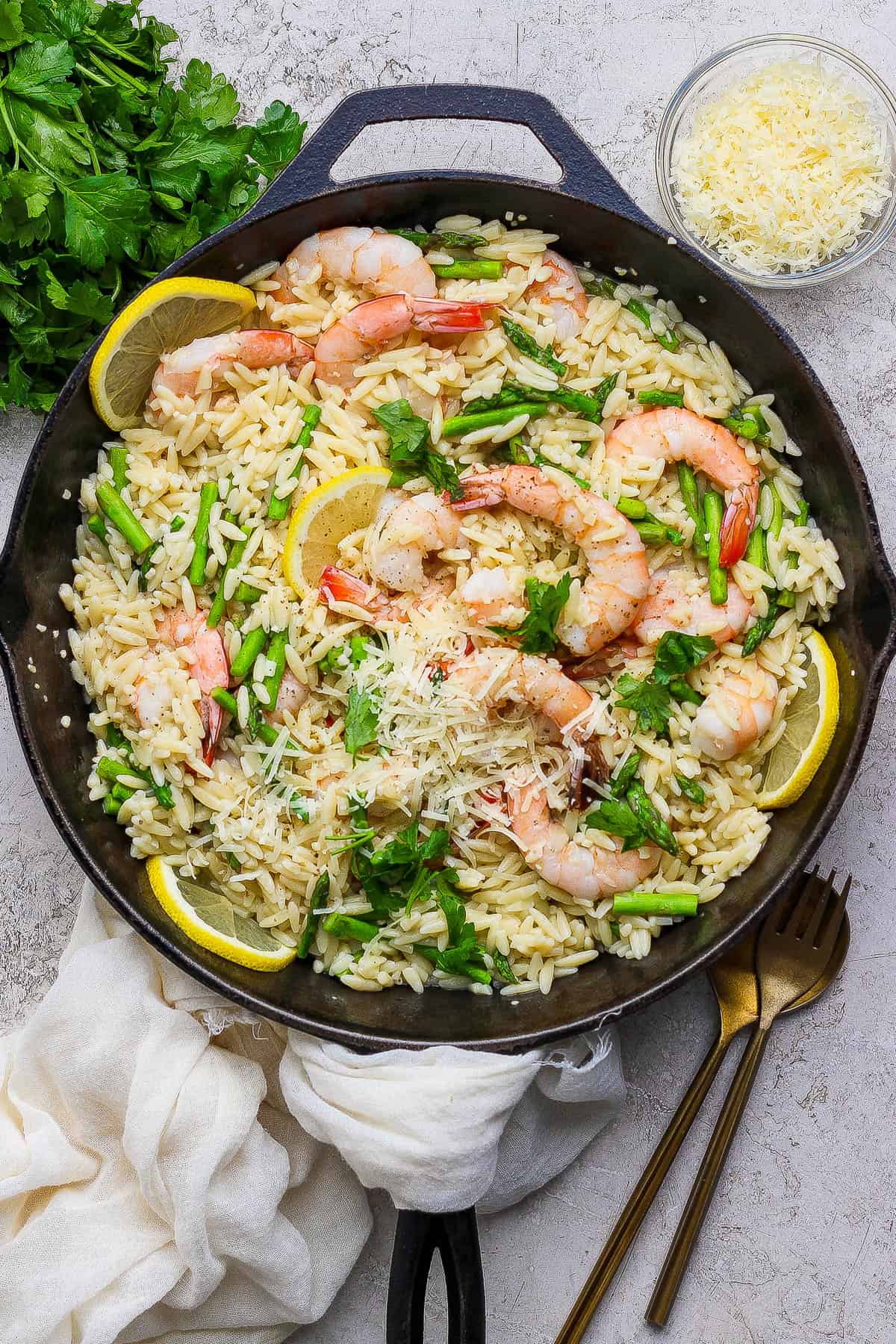 Shrimp scampi with orzo in a cast iron skillet with lemon slices next to a small bowl of parmesan and some fresh parsley.
