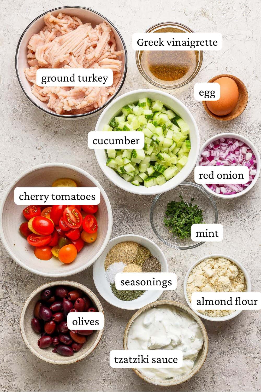 Greek turkey meatballs with orzo ingredients in individual bowls including ground turkey, greek vinaigrette, egg, cucumber, red onion, mint, cherry tomatoes, seasonings, almond flour, olives, and tzatziki sauce.