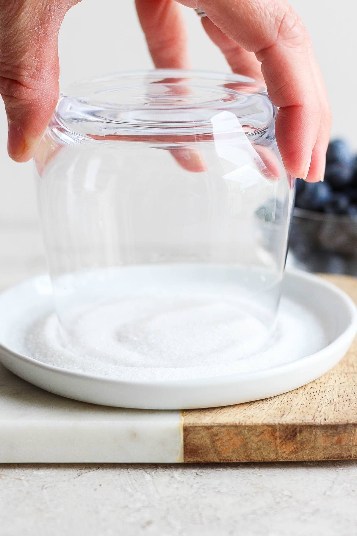 A hand holding a glass upside down in a shallow plate filled with sugar to create a sugar rim for a blueberry mojito.