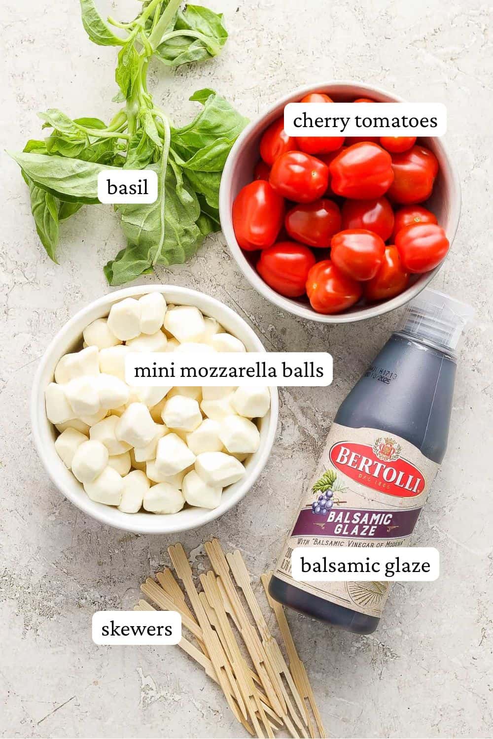 Caprese Skewers with balsamic glaze ingredients on a counter top including fresh basil, cherry tomatoes, mini mozzarella balls, balsamic glaze, and mini skewers.