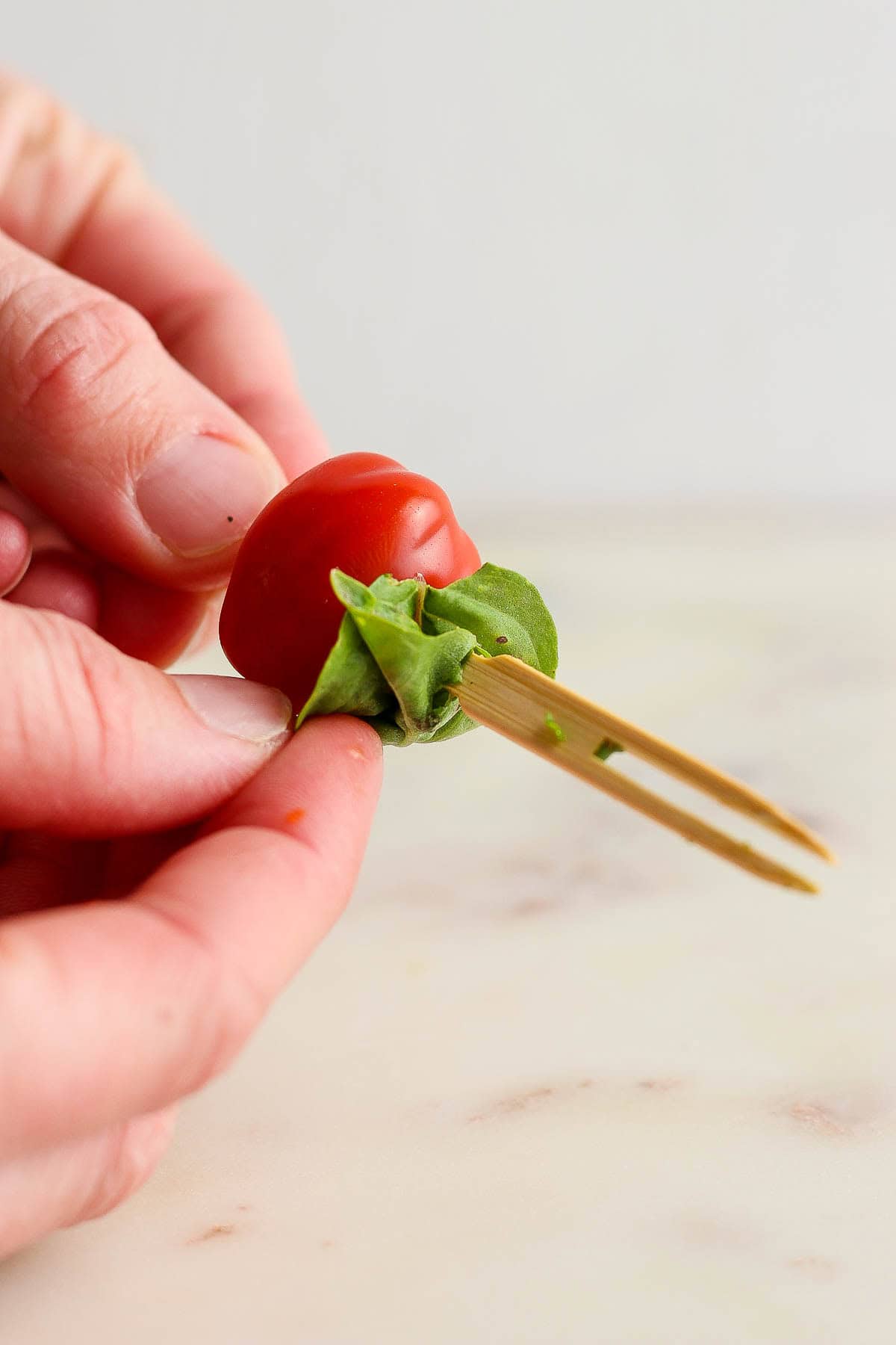 A hand holding and assembling a caprese skewer with balsamic glaze with one tomato followed by a piece of fresh basil on a skewer.