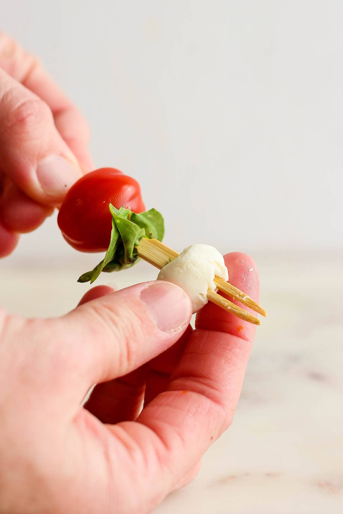 A hand holding and assembling a caprese skewer with balsamic glaze with one tomato, fresh basil, and a mozzarella pearl on a skewer.