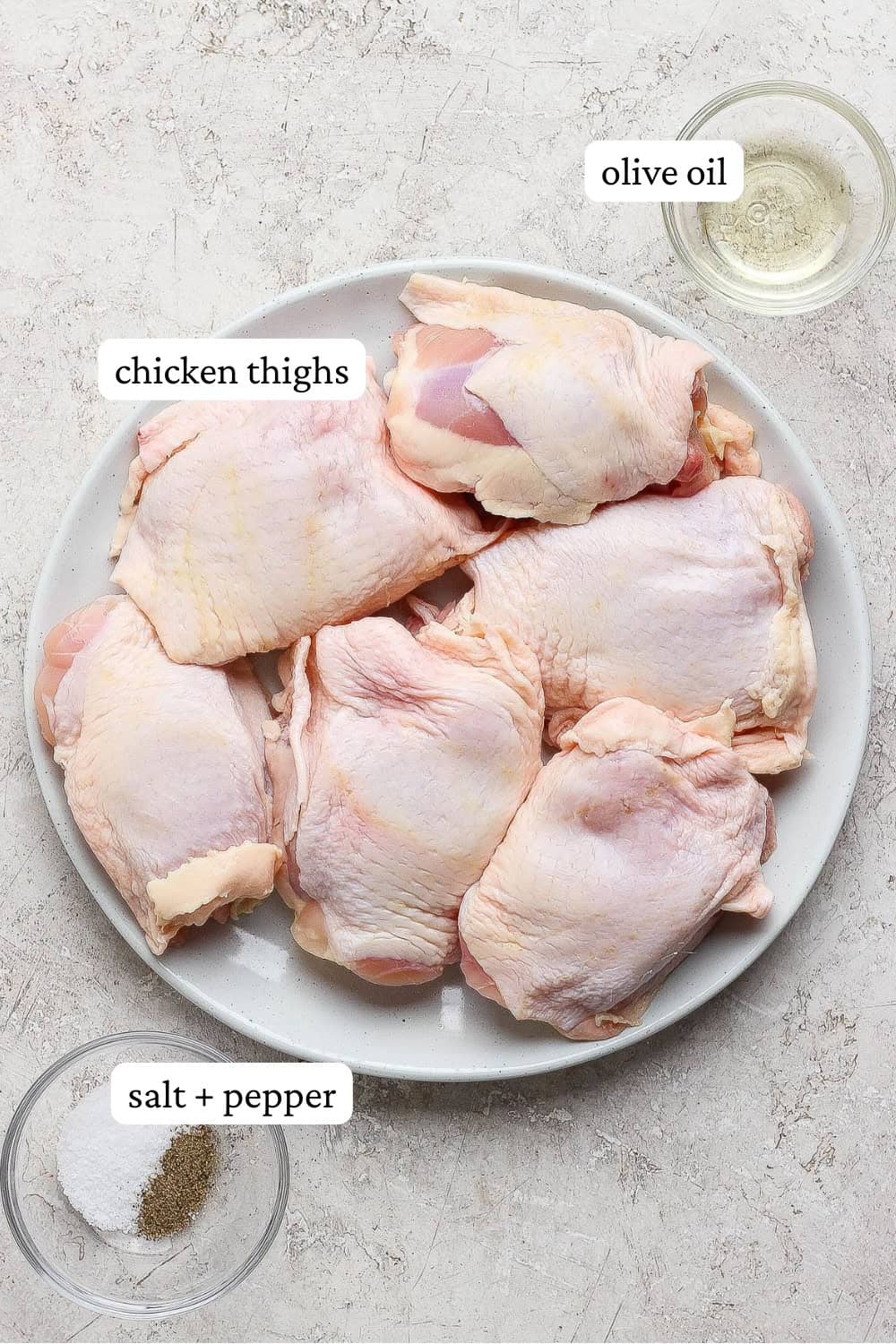 Chicken thighs on a plate next to bowls of oil and salt and pepper with labels.