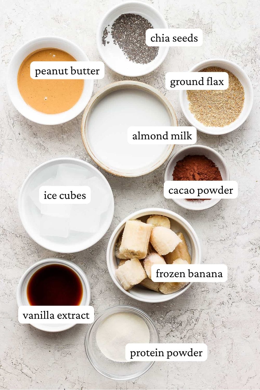 Chocolate peanut butter protein shake ingredients in individual bowls including chia seeds, peanut butter, ground flax, almond milk, ice cubes, cacao powder, frozen banana, vanilla extract, and protein powder.