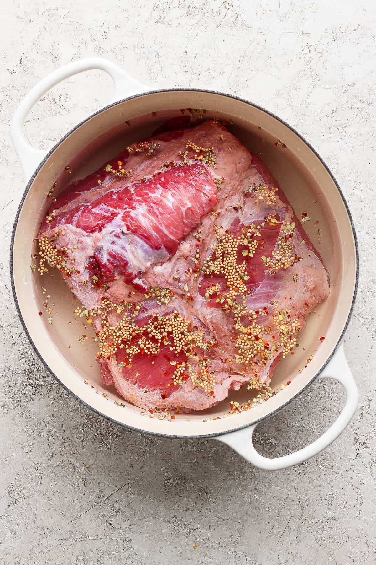 A corned beef brisket with seasoning and water in a white dutch oven before cooking.