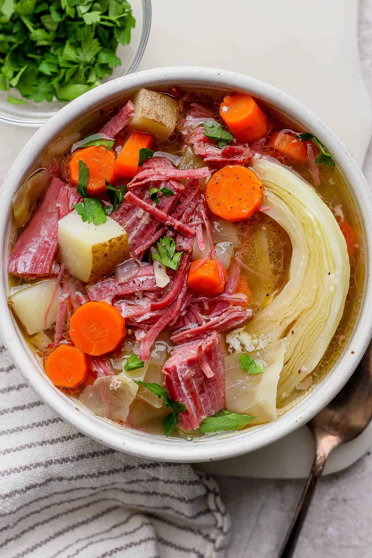 Corned beef and cabbage soup in a bowl next to a small bowl of parsley, a spoon, and kitchen towel.