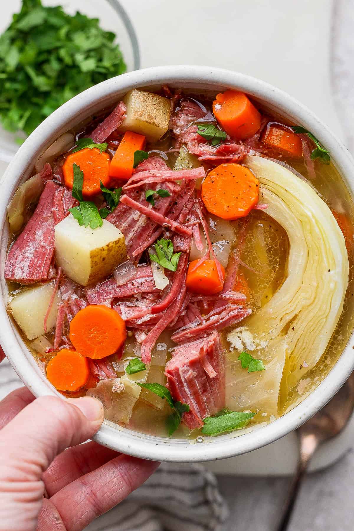 A bowl of corned beef and cabbage soup being held by a hand next to a small bowl of chopped parsley.