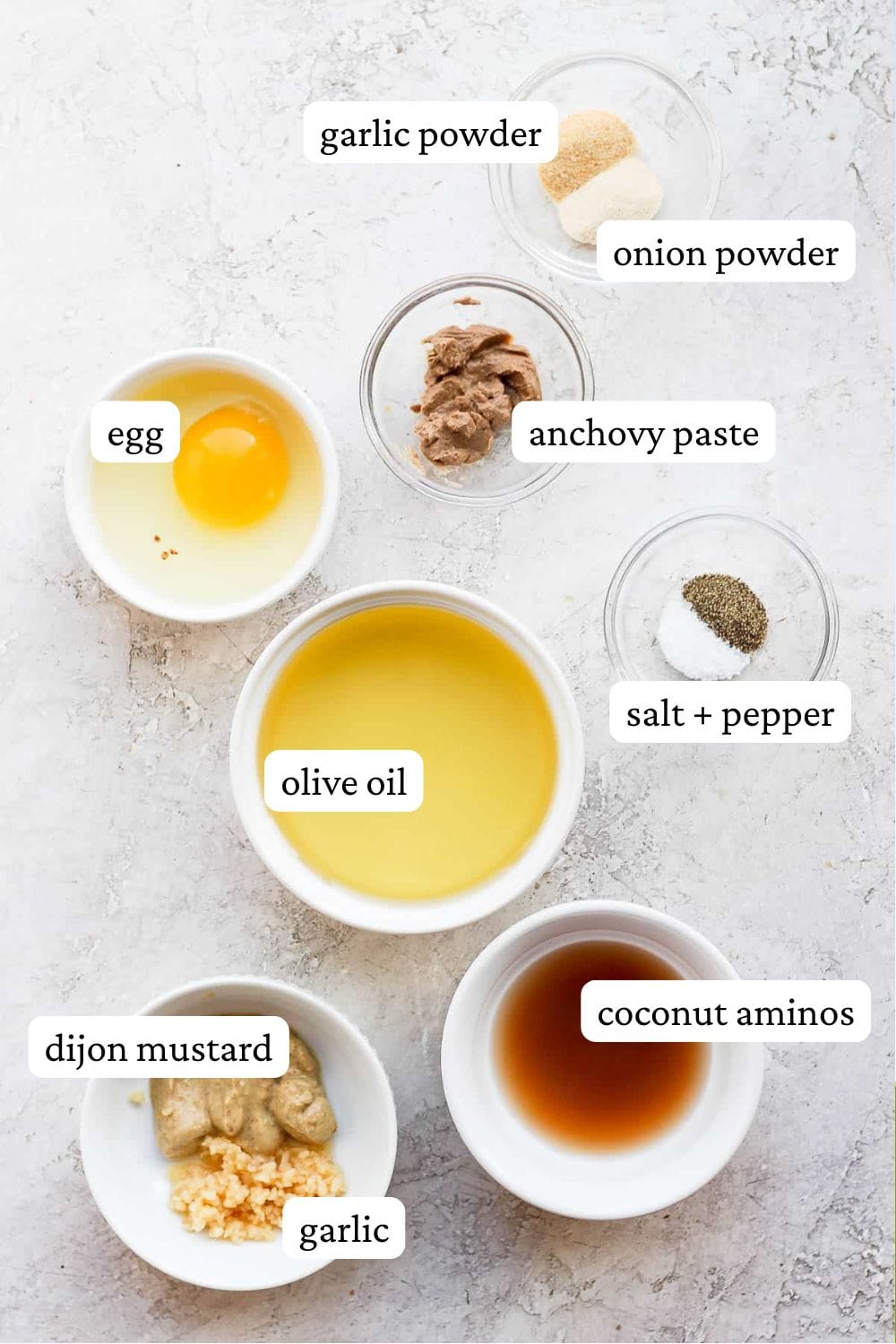 Dairy free caesar dressing ingredients in individual bowls; garlic powder, onion powder, anchovy paste, egg, salt and pepper, olive oil, dijon mustard, coconut aminos, and minced garlic.
