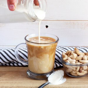 Cashew coffee creamer being poured into a glass mug of coffee next to a bowl of cashews.
