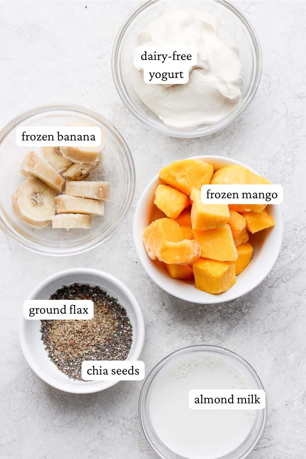 Mango banana smoothie ingredients in individual bowls including dairy-free yogurt, frozen banana, frozen mango, ground flax, chia seeds, and almond milk.