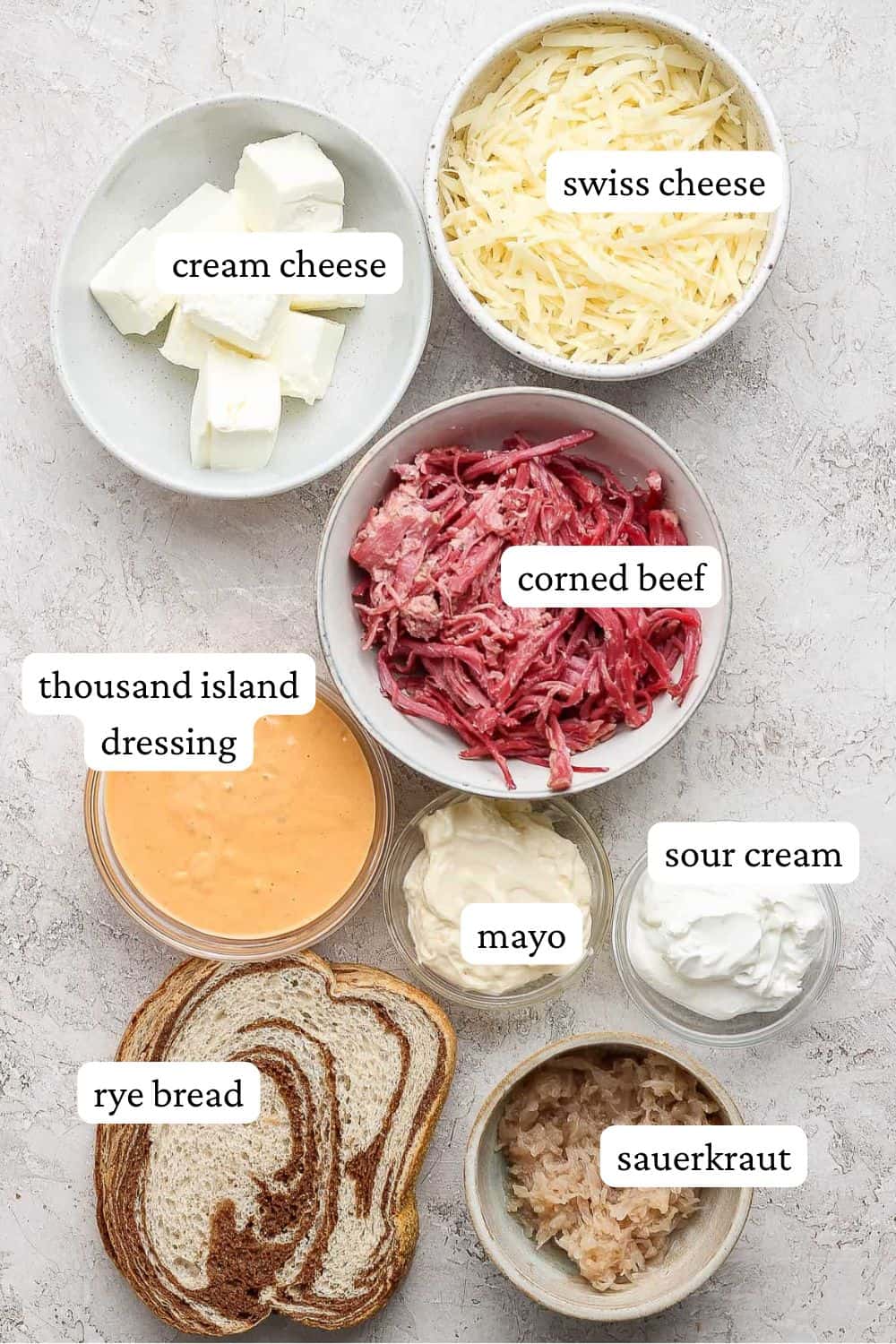 Reuben dip ingredients in individual bowls including cream cheese, shredded swiss cheese, shredded corned beef, thousand island dressing, mayo, sour cream, rye bread, and sauerkraut.