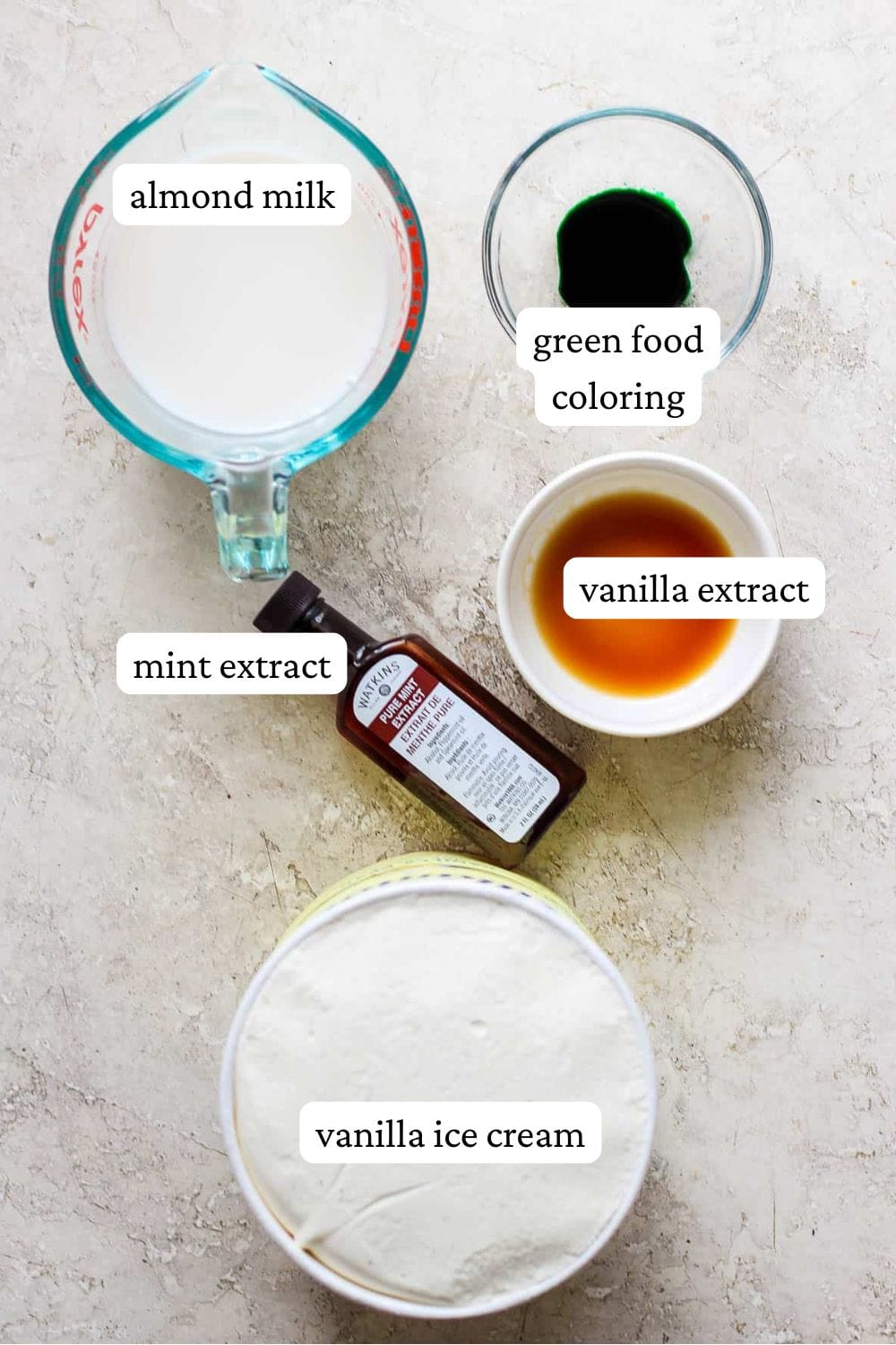 Shamrock shake ingredients in individual bowls; almond milk, green food coloring, vanilla extract, mint extract, and vanilla ice cream.