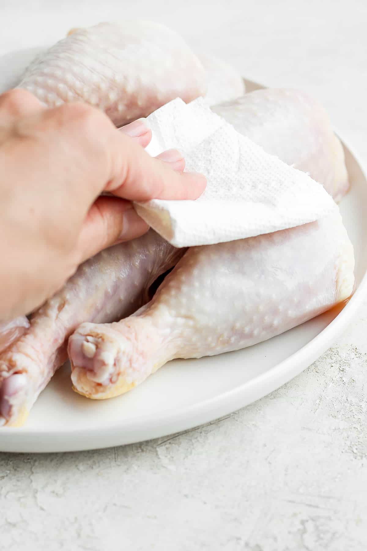 Chicken legs on a white plate being pat dry with paper towel.