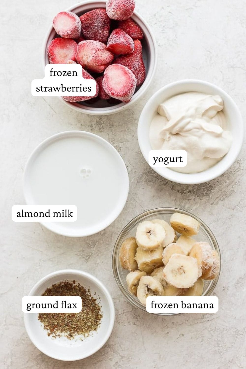 Strawberry smoothie ingredients in separate bowls including frozen strawberries, yogurt, almond milk, frozen banana, and ground flax.