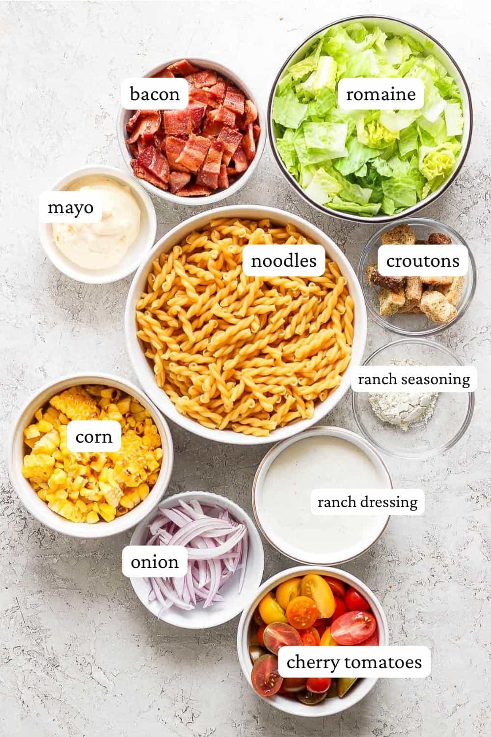 Bacon ranch pasta salad ingredients in individual bowls including cooked bacon, chopped romaine, mayo, noodles, croutons, ranch seasoning, corn, ranch dressing, red onions, and cherry tomatoes.