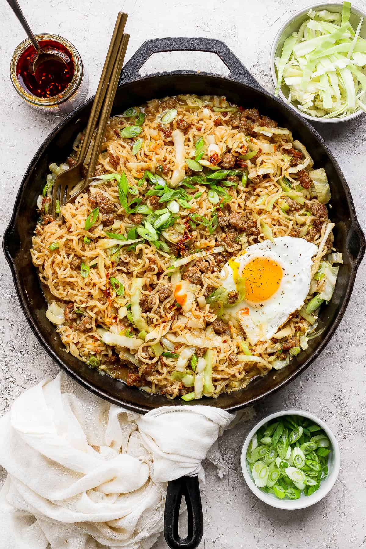 Cabbage and noodles in a cast iron skillet with an over easy egg on top next to small bowls of green onions, cabbage, and chili crisp.