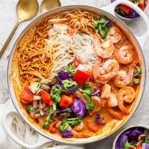 Shrimp and rice noodles with sweet curry sauce in a braising pan next to two gold spoons and small bowls of roasted vegetables.