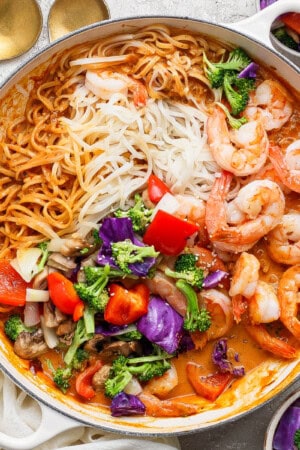 Shrimp and rice noodles with sweet curry sauce in a braising pan next to two gold spoons and small bowls of roasted vegetables.