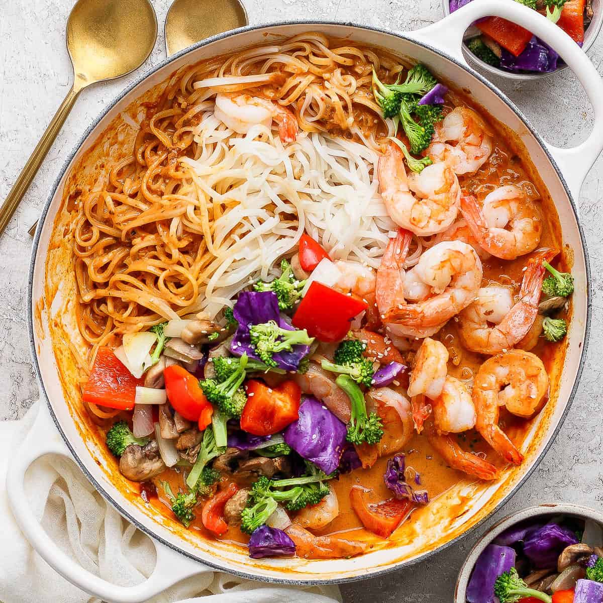 Shrimp and rice noodles with sweet curry sauce in a braising pan next to two gold spoons and small bowls of roasted vegetables.