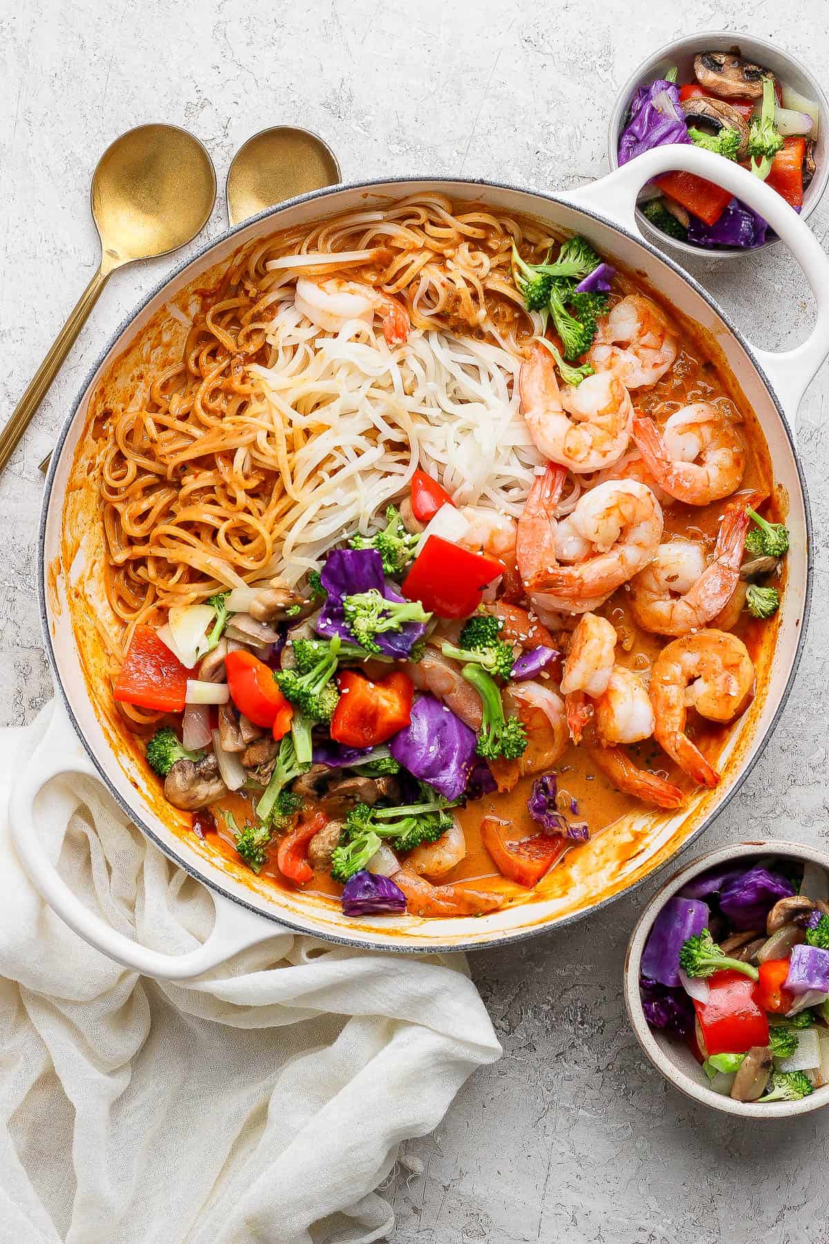 Shrimp and rice noodles in a braising pan with sweet curry sauce and roasted vegetables next to two gold spoons.