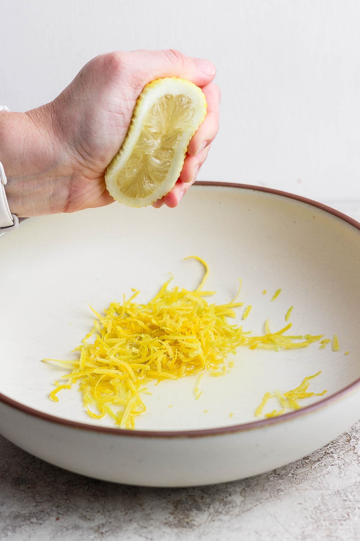 A hand squeezing half of a lemon over a serving bowl with lemon zest in it.