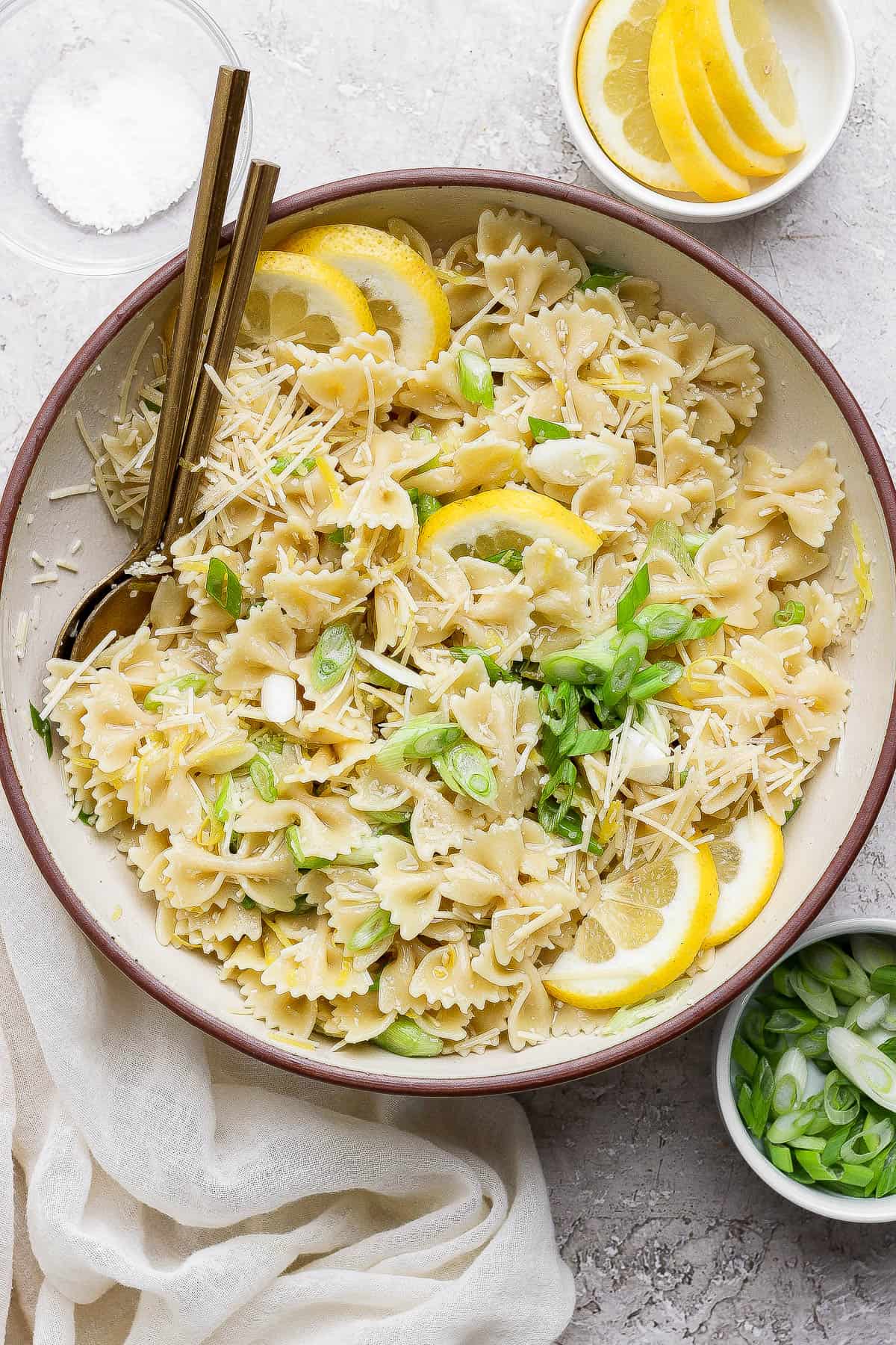 A fully mixed lemon pasta salad in a serving bowl next to small bowls of green onions, lemon slices, and salt.