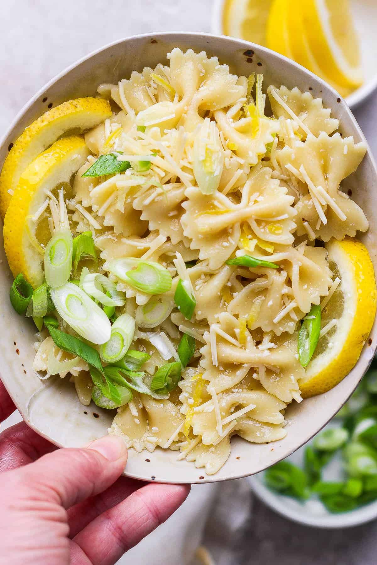 A hand holding a bowl of lemon pasta salad with lemon slices and green onions.