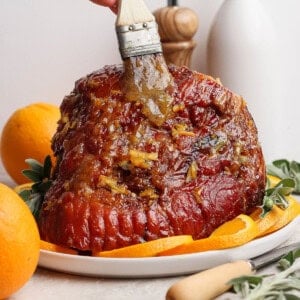 A fully cooked orange glazed ham on a platter being brushed with more glaze.