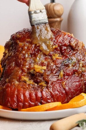 A fully cooked orange glazed ham on a platter being brushed with more glaze.