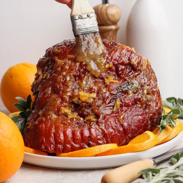 A fully cooked orange glazed ham on a platter being brushed with more glaze.
