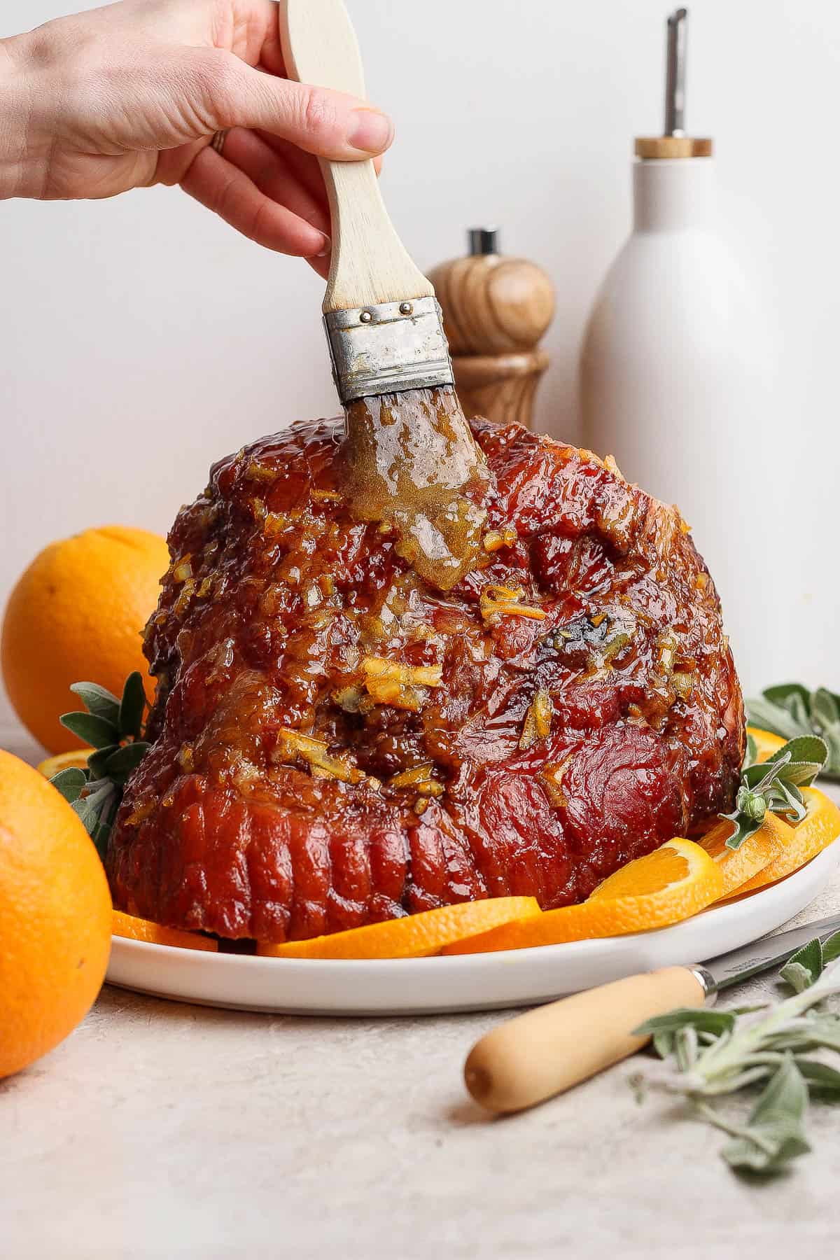 An orange glazed ham being brushed with some extra glaze before serving on a platter.
