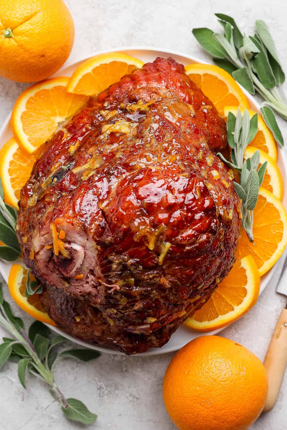 An orange glazed ham on a platter with orange slices and fresh sage.