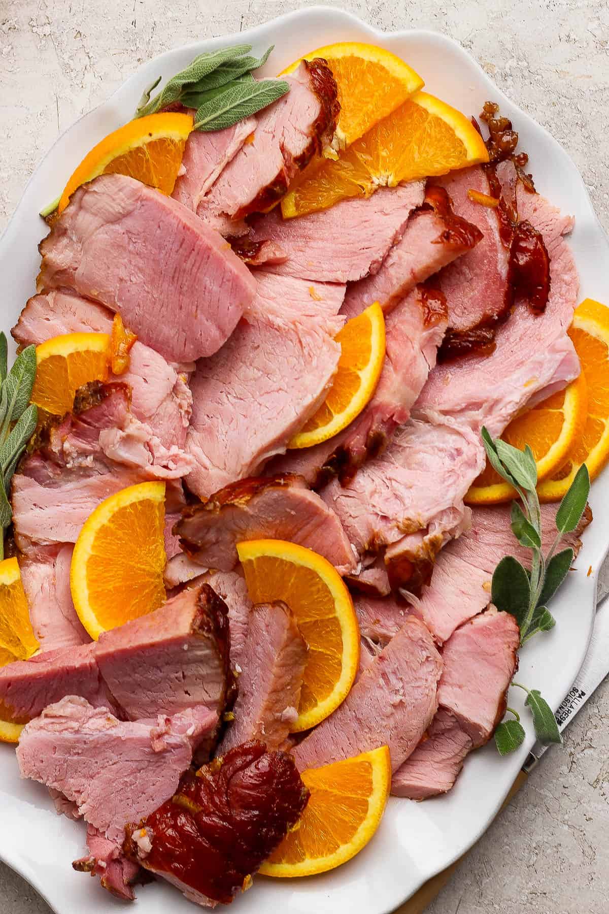 Slices of an orange glazed ham on a platter with orange slices and fresh sage.