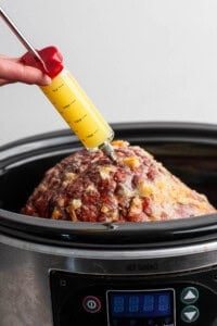 An orange glazed ham in a slow cooker being injected with orange juice.