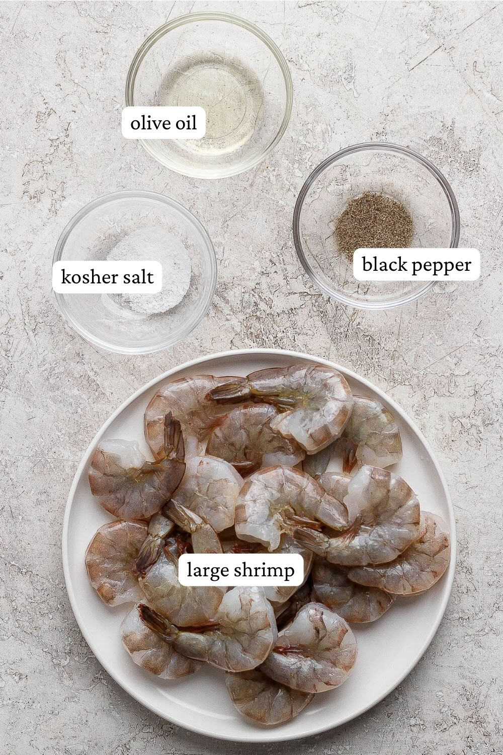 Raw shrimp on a plate next to small bowls of salt, pepper, and olive oil with labels.
