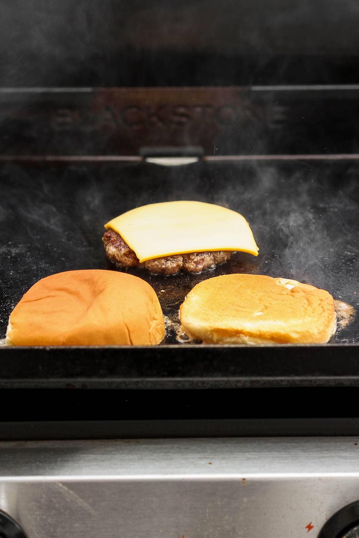 A slice of cheese added to a Blackstone smash burger with a hamburger bun toasting next to it.