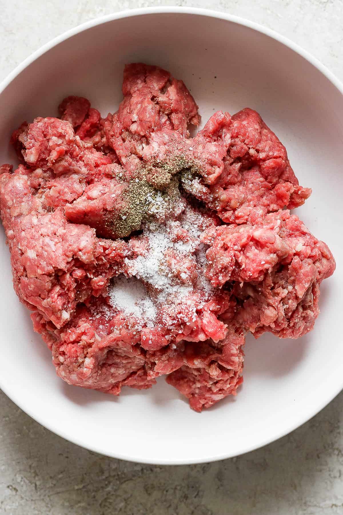 Ground beef in a white bowl with salt and pepper before being formed into smash burger balls.