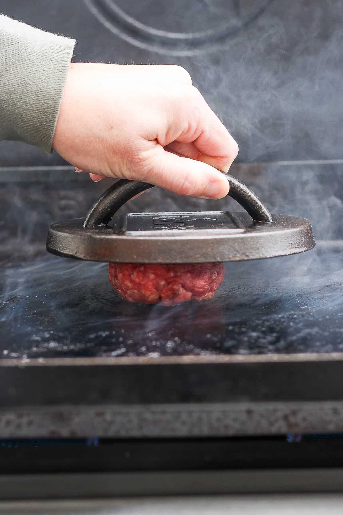 A hand using a burger press to flatten a burger ball on the blackstone.