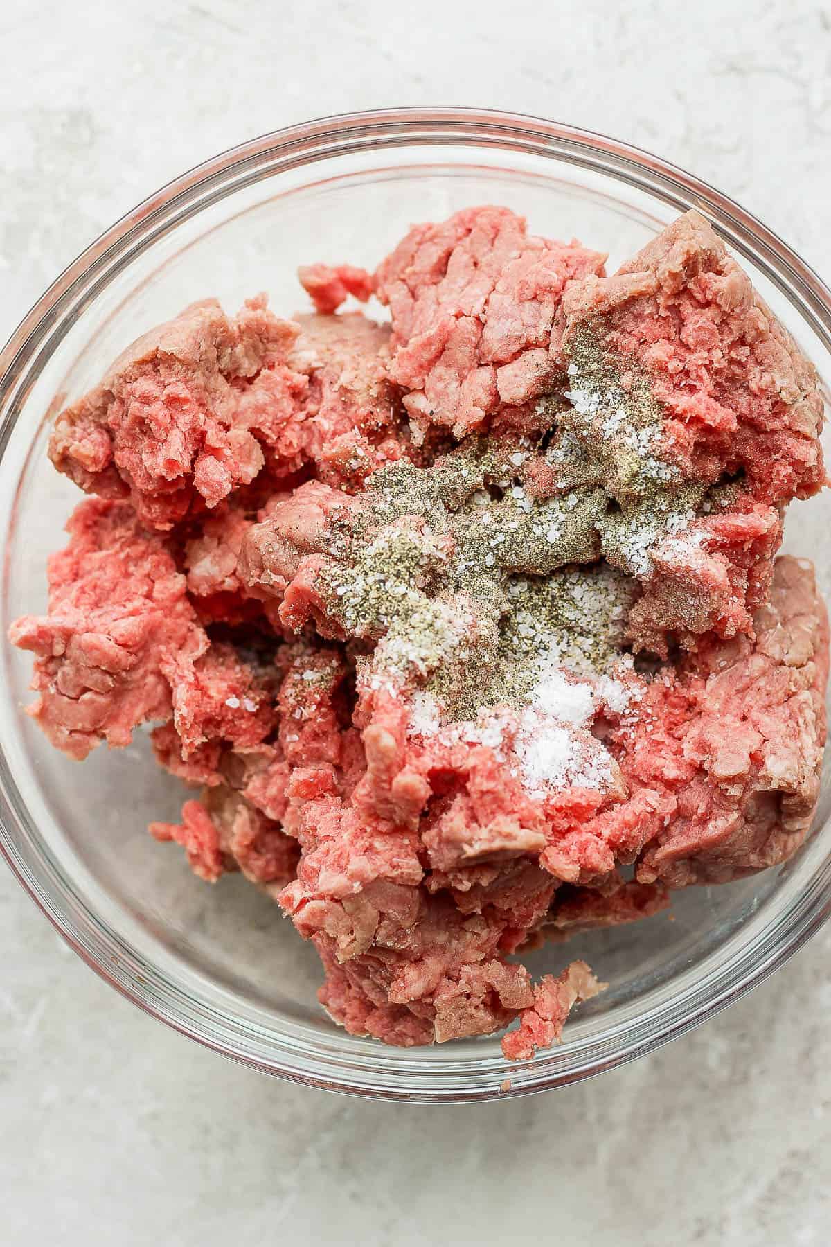 Ground beef in a large glass bowl with salt, pepper, onion powder, and garlic powder.