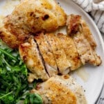 Three stove top chicken thighs on a plate next to some fresh greens.