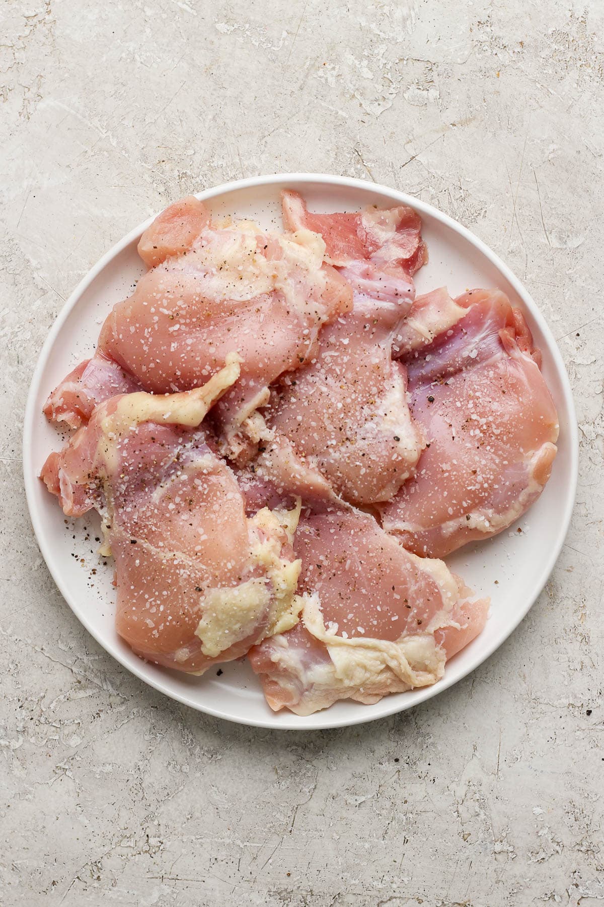 Boneless skinless chicken thighs seasoned with salt and pepper on a white plate.